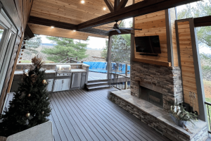 A beautifully designed outdoor living space featuring a covered deck with wood and stone finishes, complete with an outdoor kitchen and a cozy fireplace. The area is well-lit with recessed lighting and has a ceiling fan for comfort. The professional touch of C-U Under Construction, your deck contractor in Champaign, IL, is evident in the meticulous craftsmanship and attention to detail.