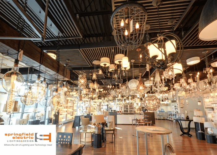 This image shows another angle of the lighting and design store interior. A variety of lighting fixtures hang from a dark, exposed ceiling structure, creating a striking visual display against the industrial backdrop. These fixtures range from elegant chandeliers to modern pendant lights and lamps. Below, the showroom floor features tables and shelves with smaller lighting products and home decor items. The store's branding "springfield electric LIGHTING & DESIGN" is again present in the lower portion of the image, alongside the slogan "Where the Art of Lighting and Technology meet." The overall lighting of the space is warm, highlighting the textures and designs of the lighting products on display. No people are visible in the scene.
