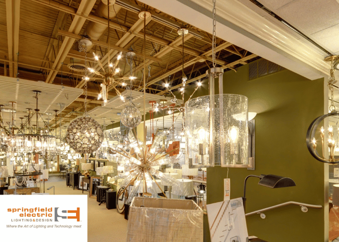This image shows the interior of a lighting and design store. Numerous light fixtures, including chandeliers, pendant lights, and lamps, are displayed throughout the space, illuminating the area with their varied designs and brightness levels. The ceiling is exposed, revealing ductwork and beams, which gives the space an industrial feel. The walls are painted in a muted green shade, and the floor seems to be covered with a beige carpet. The store's branding "springfield electric LIGHTING & DESIGN" is visible in the bottom left corner, with a tagline "Where the Art of Lighting and Technology meet." There is no visible presence of customers or staff, focusing the view on the products themselves.